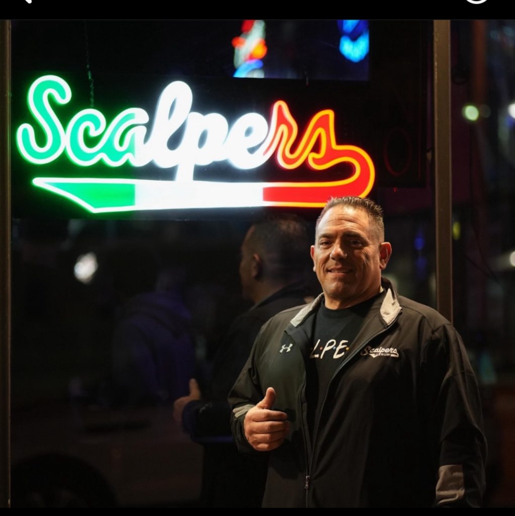 Frank Pines standing beneath the illuminated Scalpers Bar & Grille sign.