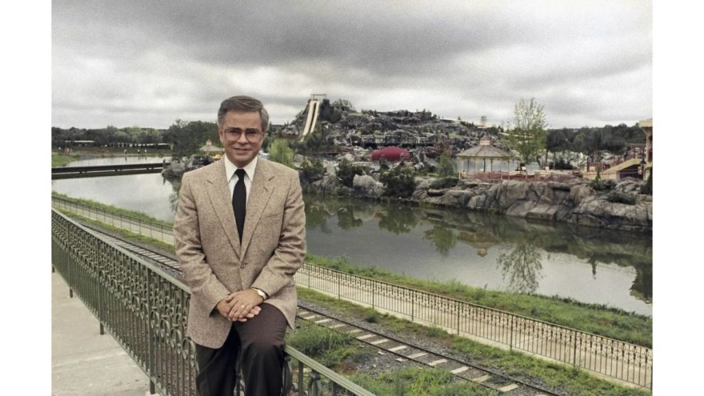 Jim Bakker standing outdoors in a tan jacket with buildings in the background.